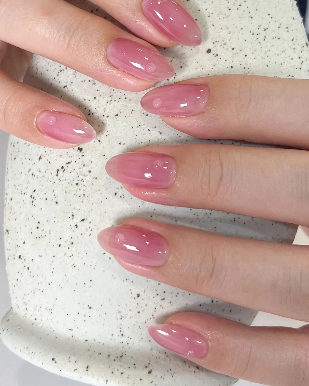 Желейний рожевий із краплинками Мигдалеподібні нігті з напівпрозорим рожевим желейним покриттям, темнішими розводами і крихітними прозорими краплинками на світлій крапчастій кераміці